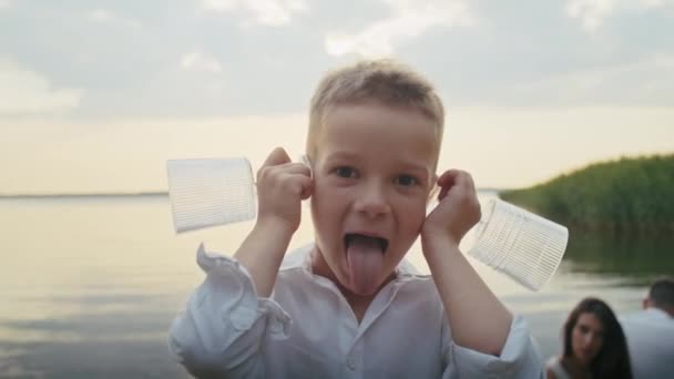 a cheerful child indulges in a pond, putting glasses to his ears and grimacing - Footage, Video