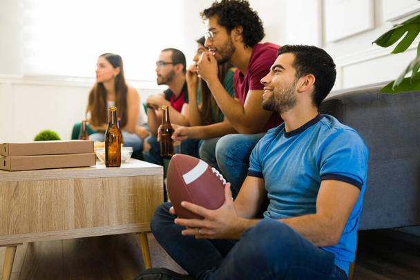 Przystojny mężczyzna trzyma piłkę nożną i uśmiecha się podczas spędzania czasu z przyjaciółmi oglądając mecz - Zdjęcie, obraz