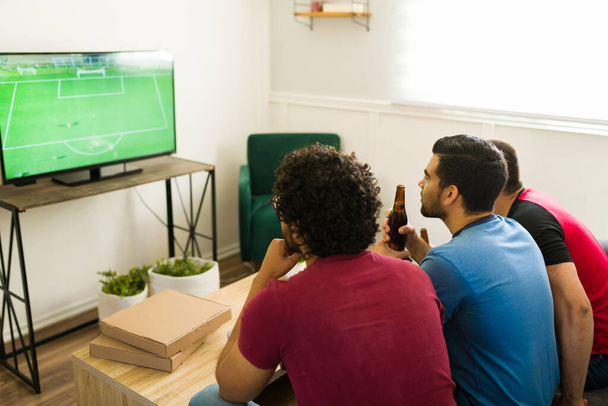 Grupo de amigos vistos desde atrás disfrutando de un partido de fútbol en la sala de estar mientras beben cerveza - Foto, Imagen