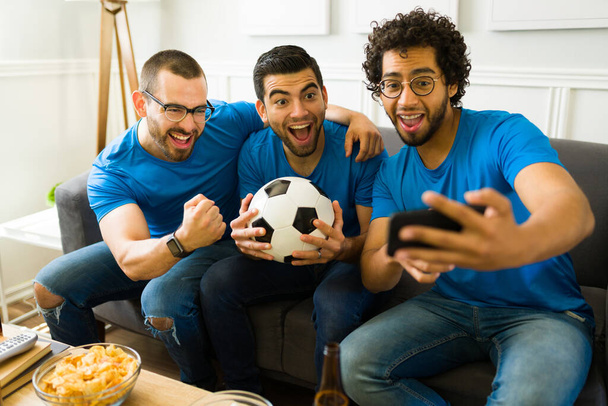 Super glückliche Gruppe von Freunden, die lachen, während sie zusammen ein Selfie machen und sich im Wohnzimmer ein Sportspiel ansehen - Foto, Bild