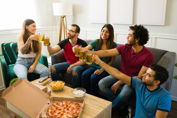 Szczęśliwi młodzi mężczyźni i kobiety wznoszący toast piwem i czujący się szczęśliwi podczas jedzenia pizzy i przekąsek w domu - Zdjęcie, obraz