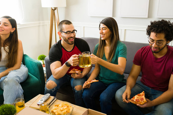 Emocionada joven pareja haciendo un brindis con cerveza y riendo mientras se divierten en una reunión de amigos - Foto, imagen