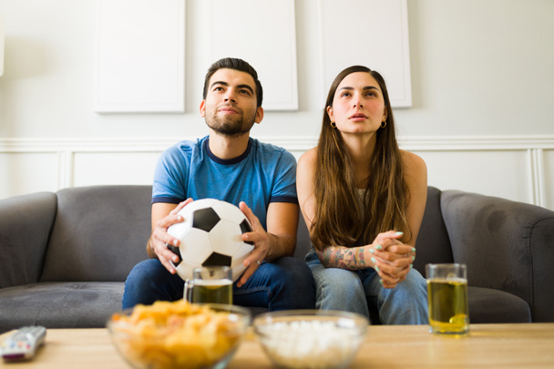 Beautiful young couple sitting on the sofa and enjoying a soccer game on TV - Photo, Image