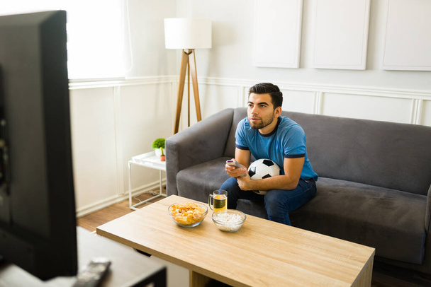 Jóképű fiatalember használja a távirányítót és nézi a focimeccset a tévében. - Fotó, kép