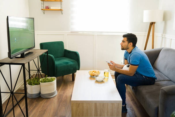 Seitenansicht eines attraktiven jungen Mannes, der sich im Fernsehen ein Fußballspiel anschaut und dabei Bier und Snacks genießt - Foto, Bild