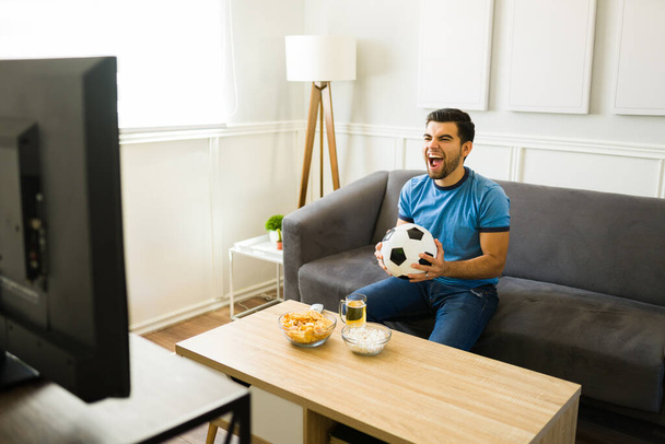 Латиноамериканець, який радіє національній команді під час гри на ТВ, сидячи на дивані. - Фото, зображення