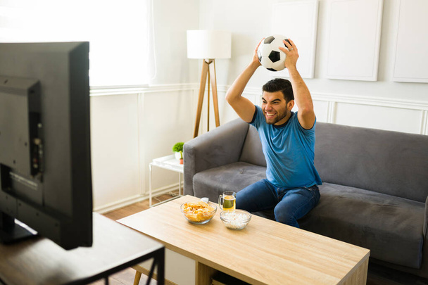 Young man feeling very excited while holding a soccer ball and watching the sports game in the living room - Photo, Image
