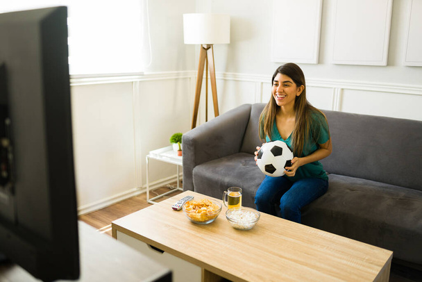 Mujer joven hispana sonriendo mientras disfruta viendo la selección nacional de fútbol en la televisión - Foto, Imagen