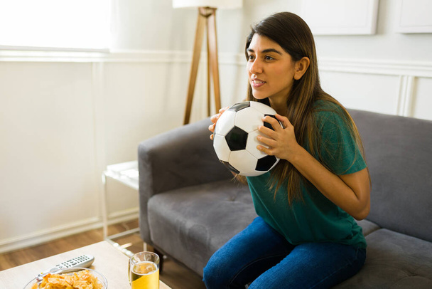 Ανησυχούσα νεαρή γυναίκα κρατώντας μια μπάλα ποδοσφαίρου και δίνοντας μεγάλη προσοχή στο αθλητικό παιχνίδι στην τηλεόραση - Φωτογραφία, εικόνα