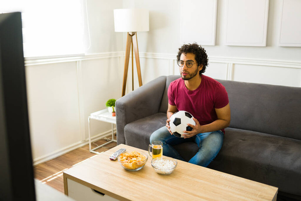 Ελκυστικός Λατίνος που κρατάει μια μπάλα ποδοσφαίρου και δίνει μεγάλη προσοχή στο αθλητικό παιχνίδι στην τηλεόραση - Φωτογραφία, εικόνα