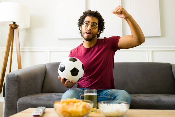 Привлекательный молодой человек в гостиной болеет за свою любимую футбольную команду после победы в чемпионате - Фото, изображение