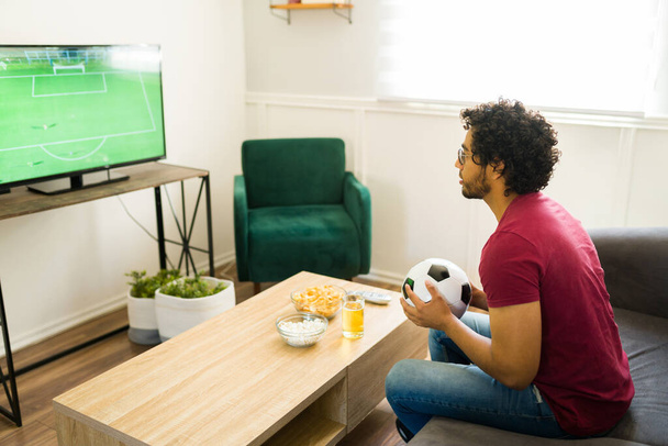 Latin sports fan holding a soccer ball while watching the championship game on television - Foto, imagen