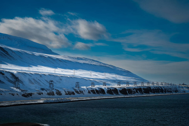 SVALBARD COASTLINE WITH OLD MINING TOWERS TO MOVE COAL ALONG A SNOW COVERED MOUNTAIN AND A LIGHT BLUE SKY - Fotó, kép