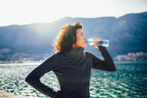 Fitness-Frau trinkt Wasser aus der Flasche. Muskulöse junge Frau macht eine Trainingspause im Freien. - Foto, Bild