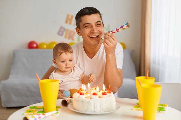 Image of young adult man with toddler baby celebrating first birthday, sitting at table, looking at birthday cake, father holding party horn, laughing, posing with his charming cute kid. - Foto, immagini