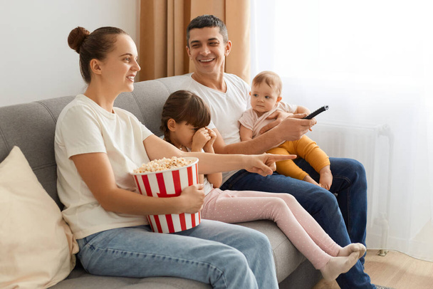 Side view of happy family watching television while eating popcorn together on the sofa, mother holding remote choosing movie or cartoon to watch, posing near window in living room. - Foto, Imagen