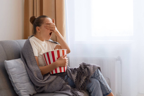 Indoor shot of shocked astonished woman with bun hairstyle wearing white t shirts wrapped in blanket watching horror movie, covering eyes with palm, keeps mouth moment. - 写真・画像