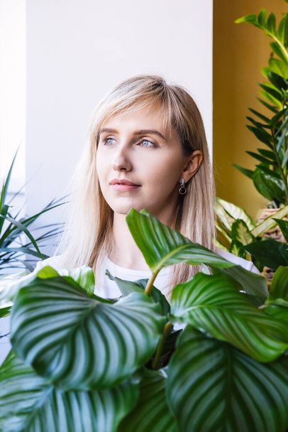 Portrait of a young blonde woman in a white t-shirt who holds big house plant calathea with a large green leaves. Vertical photo. - Foto, afbeelding
