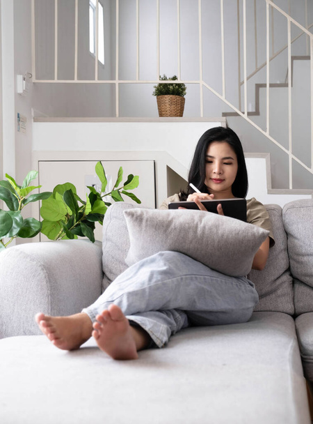 Приваблива усміхнена молода азіатка, що розслабляється на шкіряному дивані вдома, працює на ноутбуці
 - Фото, зображення