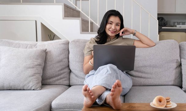 Piękna uśmiechnięta Azjatka patrząca na kamerę relaksująca się na skórzanej sofie w domu. praca na laptopie - Zdjęcie, obraz