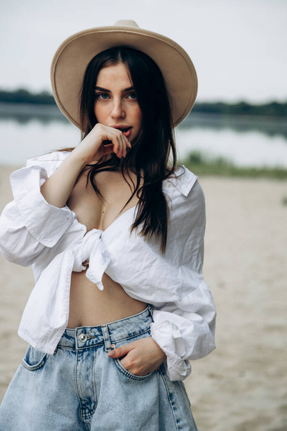 白いシャツと水着で日焼けしたセクシーな体を持つ幸せな若い女の子の帽子を持つ美しいブロンドの女性の夏のライフスタイルの肖像画は、澄んだ水と熱帯の島のビーチに立っています - 写真・画像