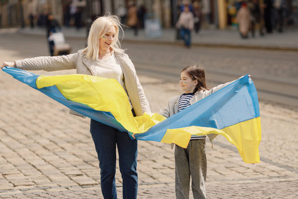 若い母親と娘が古い町の通りに立っている。女の子と女性は青と黄色の色でウクライナの大きなフラグを保持します。戦争に対するウクライナを支援する女性と少女. - 写真・画像