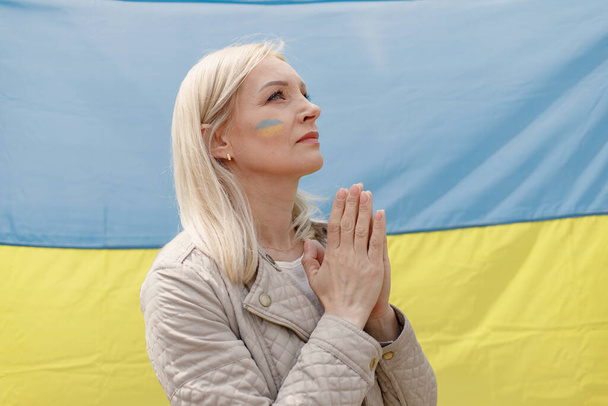 Woman with ukrainian flag on a cheek standing in front of big flag of Ukraine. Girl praying. Girl supporting Ukraine. - Fotoğraf, Görsel