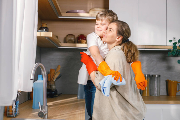 Blonde boy and adult woman cleaning in a kitchen and wearing orange gloves. Caucasian boy helping his mother to do housework. Mother hugging her son. - Foto, Imagem