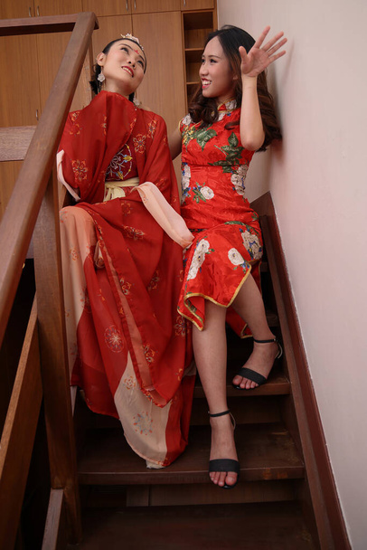 dois jovem atraente mulher asiática vestindo tadeonal chinês vermelho hanfu saia longa cachecol e cheongsam vestido de fantasia sentar na escada de madeira interior pose falar - Foto, Imagem