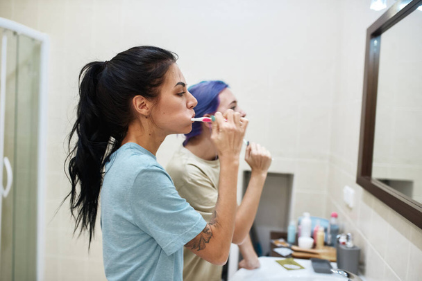 Side view of young lesbian caucasian girls brushing teeth with toothbrushes at morning. Domestic lifestyle. Hygiene and dental care. Persons looking on their reflection in mirror. Modern bathroom - Fotografie, Obrázek