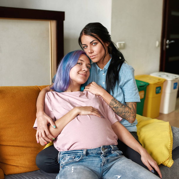 Girl hug her pregnant girlfriend partner on sofa at home. Young caucasian lesbain couple look at camera and spend time together. Homosexual relationship and motherhood. Closeness. Domestic lifestyle - Foto, imagen