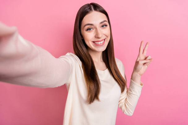 Self-portrait of attractive cheerful brown-haired girl blogger showing v-sign isolated over pink pastel color background. - Photo, image