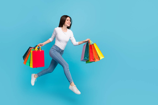 Photo of adorable dreamy woman wear white shirt jumping high running holding bargains isolated blue color background. - Foto, immagini
