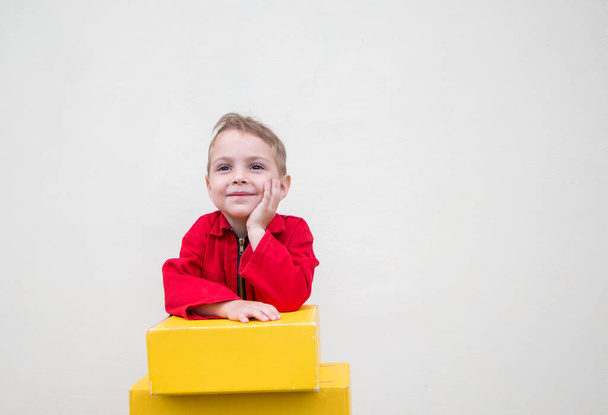 3-4 éves, boldog arc egy piros overálos fiútól, sárga kartondobozok közelében, csomagoknak. Küldemény, kis postás. Dolgozz úgy, mint egy apa. pozitív visszajelzés. reklámszöveg helye - Fotó, kép