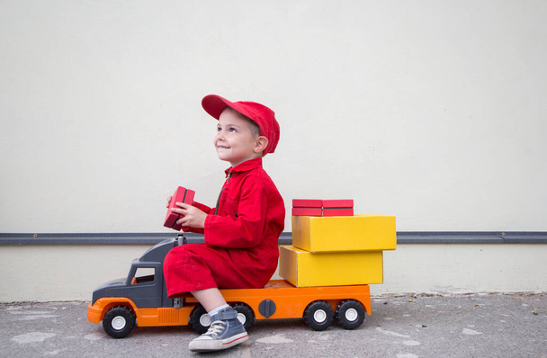 милый улыбающийся 3-4-летний мальчик в красном комбинезоне униформе и кепке сидит на большой игрушечной машине грузовик с картонными коробками посылок. Помощник почтальона, водитель грузовика. Бизнес-реклама - Фото, изображение