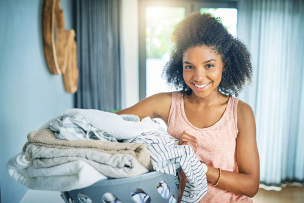 Portrait of a cheerful young woman holding a basket of washing while looking at the camera at home during the day. - Foto, imagen
