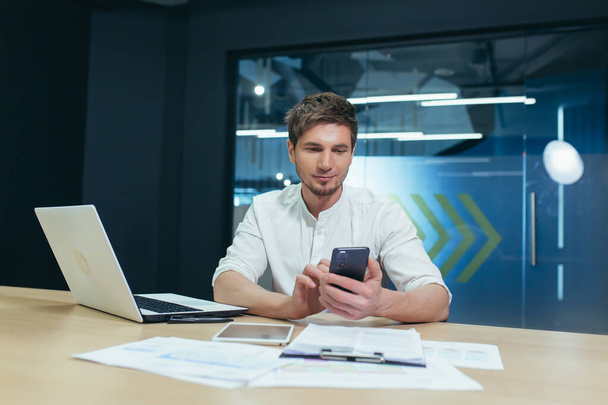 Молодой бизнесмен с бородой работает в современном офисе, с ноутбуком смотрит на экран телефона, улыбается читает хорошие новости - Фото, изображение