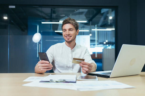Young man in office working with laptop uses phone and bank credit card for online shopping in online store, happy smiling businessman - Fotografie, Obrázek