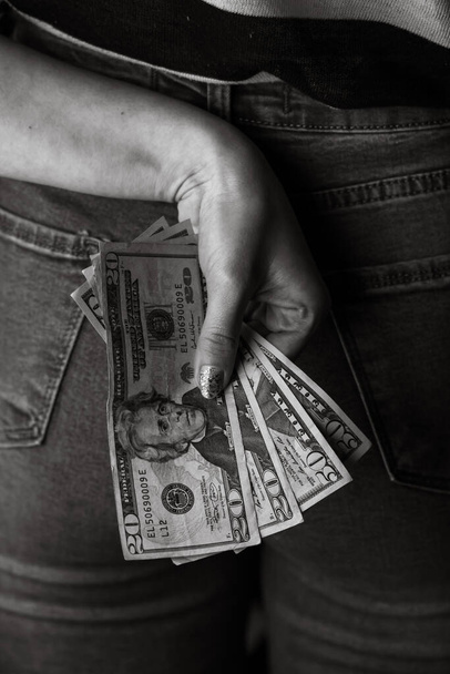 Mano de mujer sacando dinero del bolsillo trasero de jeans. Mujer escondiendo dinero a sus espaldas. Billetes en dólares en primer plano
 - Foto, Imagen