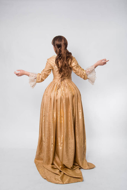 Full length portrait of a woman in a gold dress in the style of the rococo era, standing with her back forward and posing isolated on a white background. - Zdjęcie, obraz