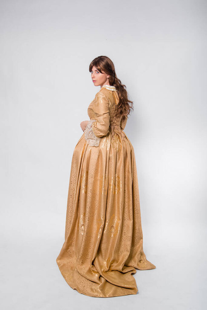 Full length portrait of a woman in a gold dress in the style of the rococo era, standing with her back forward and posing isolated on a white background. - Photo, image