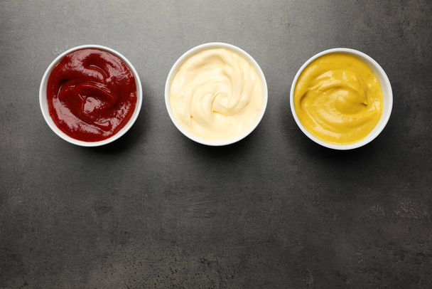 Ketchup, mustard and mayonnaise in bowls on grey table, flat lay. Space for text - Valokuva, kuva