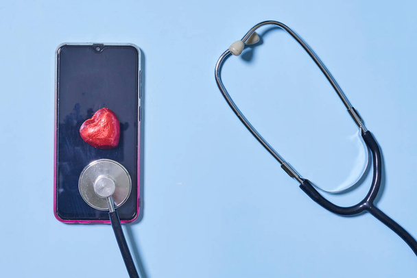 青い背景に聴診器、スマートフォン、赤いハート。医療、医療、疾病予防のテーマ. - 写真・画像