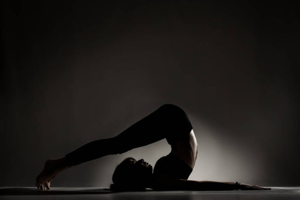 Fit ballerina girl lying and stretching practicing yoga poses. Side lit silhouette against dark background. - Foto, immagini