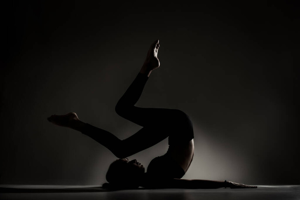 Fit ballerina girl lying and stretching practicing yoga poses. Side lit silhouette against dark background. - Valokuva, kuva