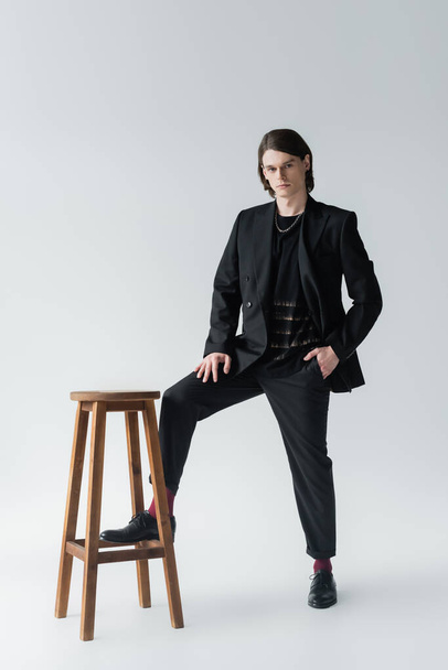 Full length of stylish man in suit posing near chair on grey background - Foto, immagini