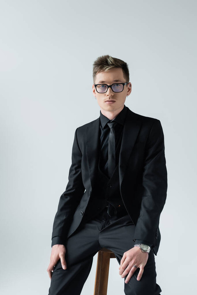 Young man in formal wear in eyeglasses sitting on chair isolated on grey  - 写真・画像