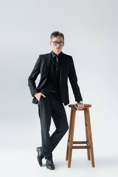 Stylish man in suit and eyeglasses posing near chair isolated on grey  - Фото, зображення
