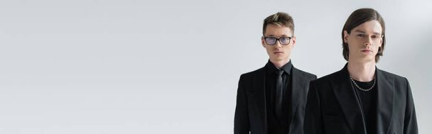 Young gay looking at camera near boyfriend in formal wear and eyeglasses isolated on grey, banner  - Fotó, kép