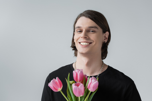 Positive young man holding pink tulips isolated on grey  - Фото, изображение